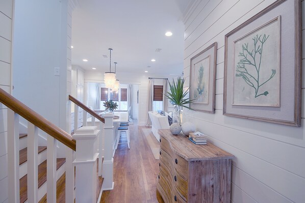 A hallway with wooden flooring, white walls, and a staircase with wooden railings. There are framed botanical prints on the wall and a wooden cabinet with decorative items.