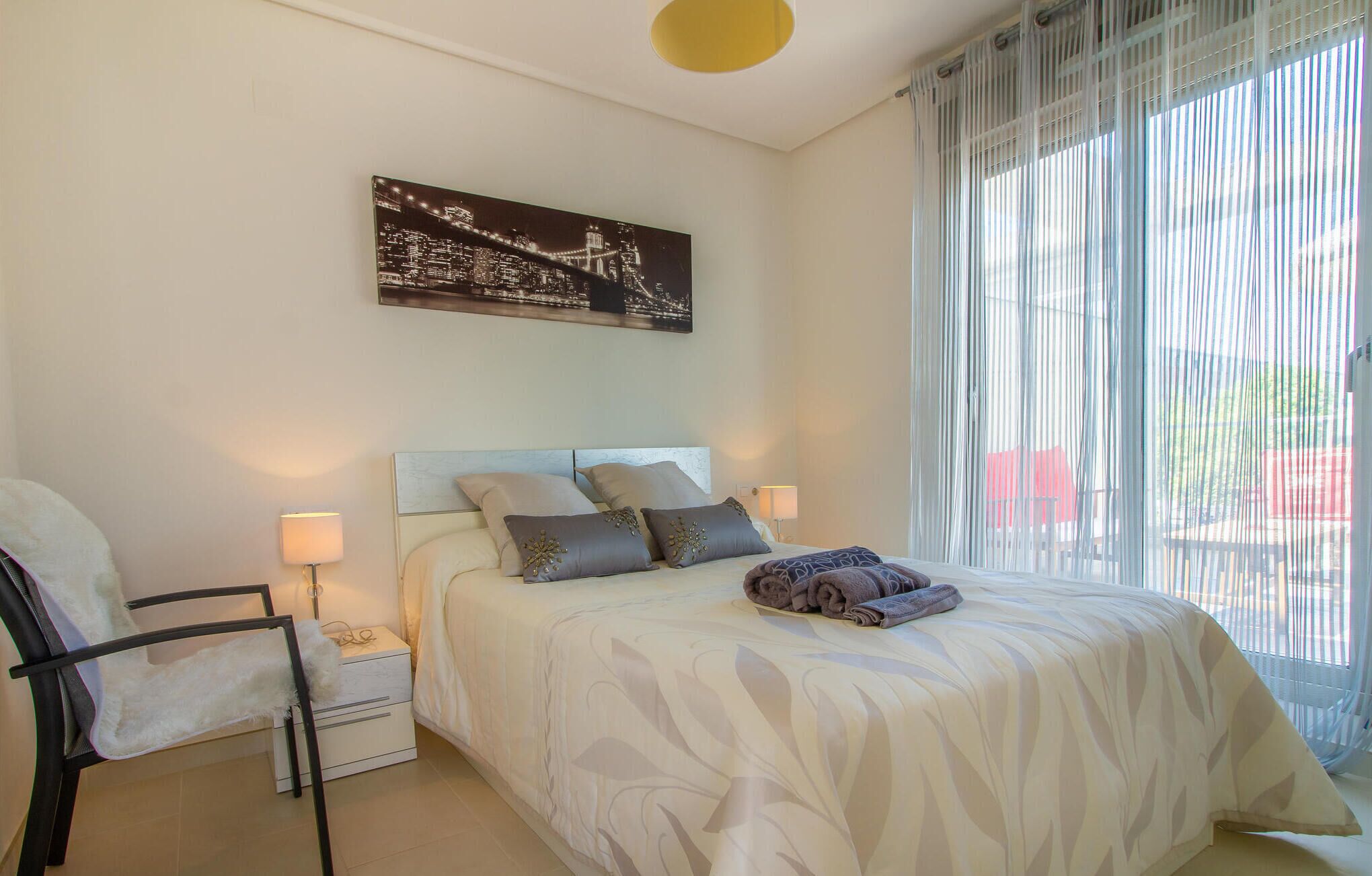 Ein Schlafzimmer mit einem Bett, einem Sessel, einem Nachttisch, einer Lampe und einem Fenster mit Vorhängen.