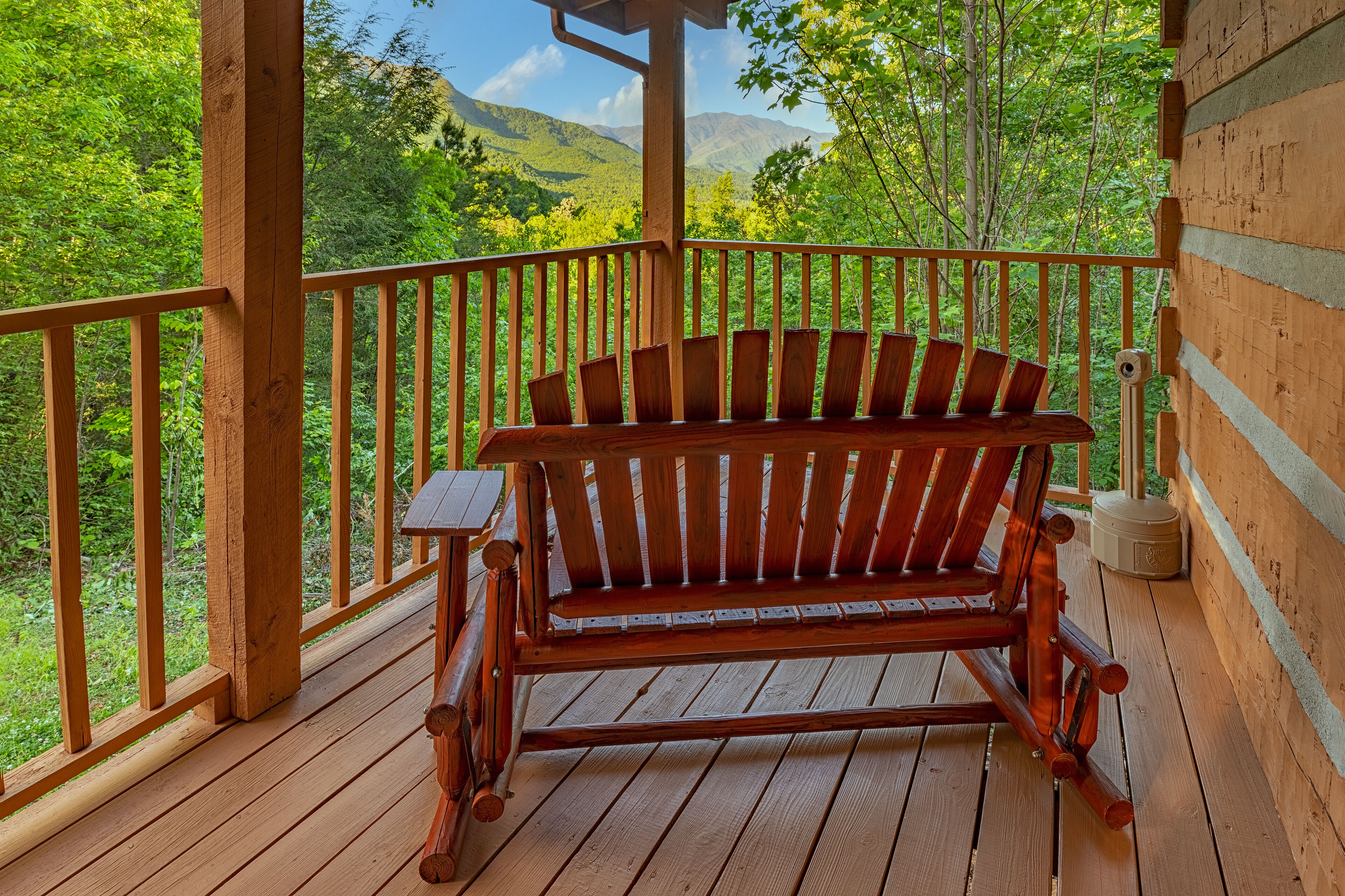 A wooden deck with a bench and a small table, overlooking a forested area with mountains in the background.