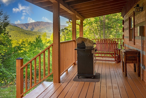 A wooden deck with a grill and chairs overlooking a forested valley.