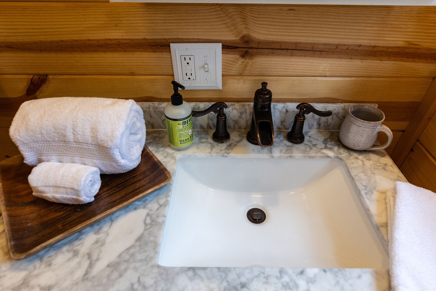 Een badkamer met een witte wasbak, houten lambrisering, een zeepdispenser en een handdoek op een houten dienblad.