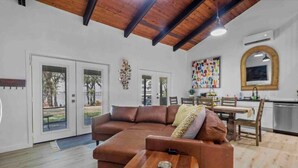 A spacious living area with a brown leather sofa, a wooden coffee table, and a dining table with chairs. There is a kitchen area with a large window and a colorful abstract painting on the wall. - Modern Lakefront Loft Retreat ~ Private Beach & Water Toys (Little Elm)