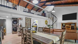 A spacious living area with a dining table, wooden chairs, a leather sofa, a staircase, and a TV mounted on the wall. - Modern Lakefront Loft Retreat ~ Private Beach & Water Toys (Little Elm)