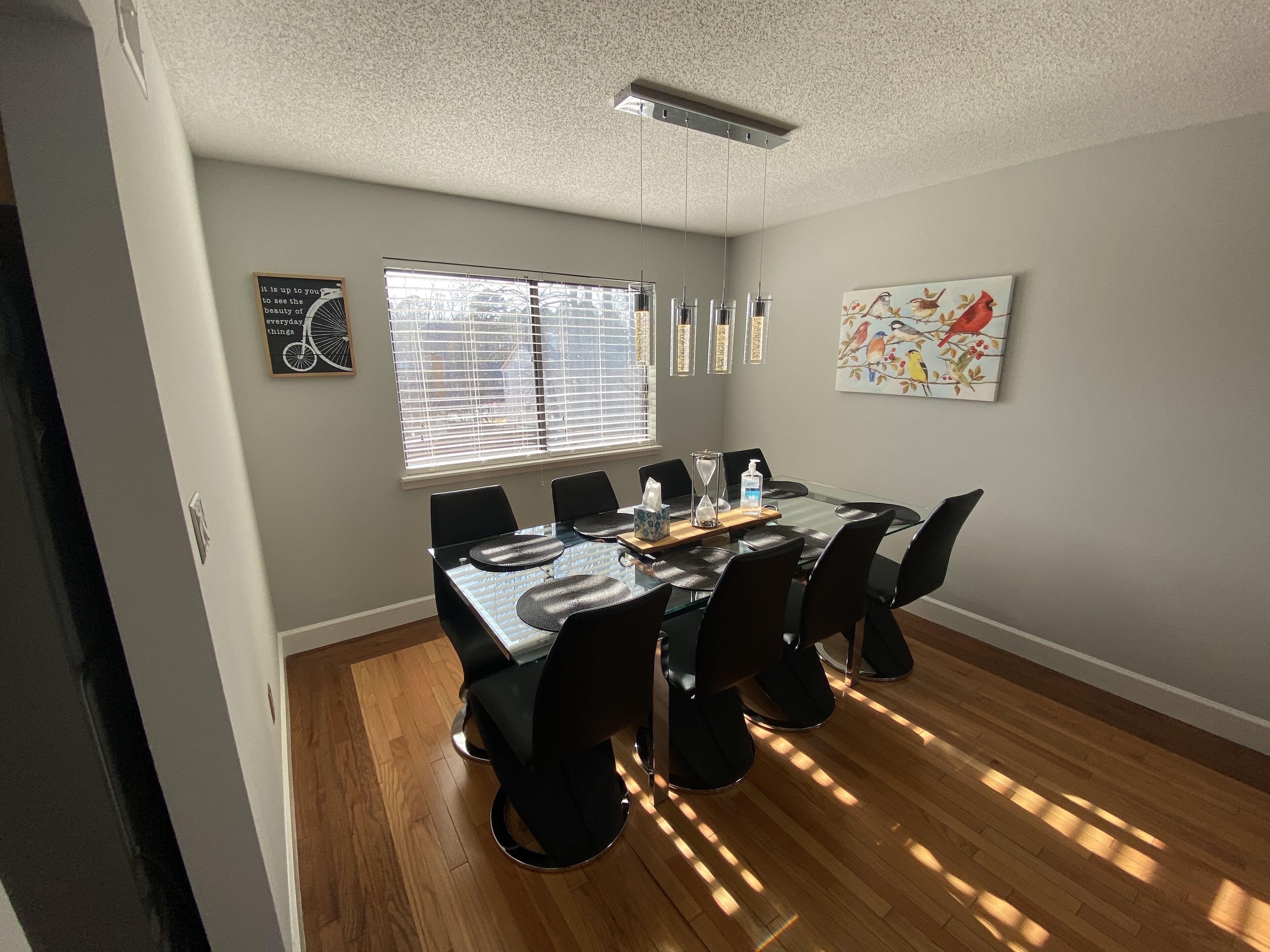 A dining area with a glass table, black chairs, a window with blinds, and a ceiling fan.