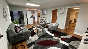 Una sala de estar moderna con sofás grises, una mesa de centro y una alfombra estampada. Se ve una cocina al fondo, con un refrigerador y un microondas.