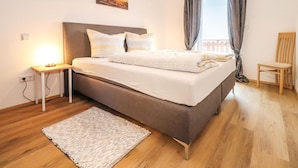 A neatly arranged bedroom with a bed, bedside table, lamp, and a view of the outdoors through a window.