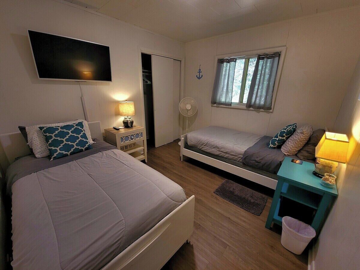 A bedroom with two beds, a TV, a window with curtains, a fan, and a small table with a lamp.