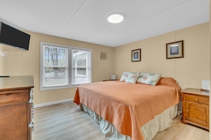 A bedroom with a bed, a dresser, a nightstand, a TV, and a window with blinds.
