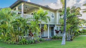 Una casa de dos pisos con un porche, rodeada de exuberantes plantas tropicales.