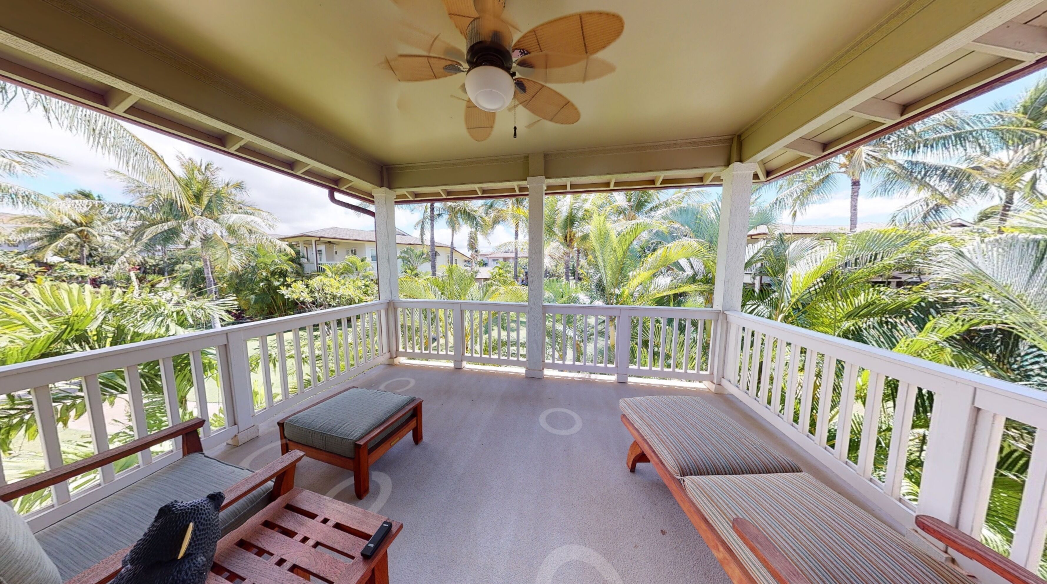 A covered outdoor area with a ceiling fan, wooden furniture, and a view of palm trees and houses.