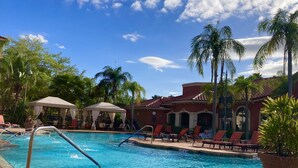 Zona de piscina con sillas de descanso y sombrillas, rodeada de palmeras y un edificio con tejado de tejas rojas.