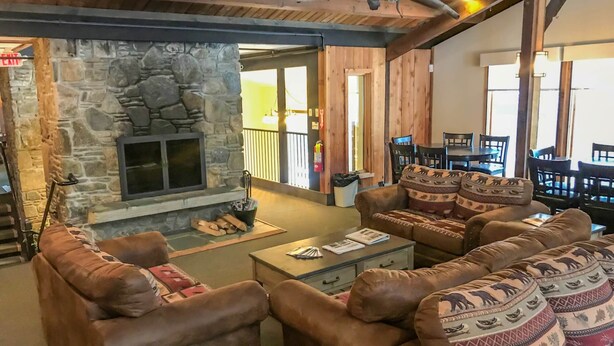 A cozy living room with a stone fireplace, leather sofas, and a dining area with wooden furniture. - Winter ‘27 Seasonal Available-Dog Friendly Chalet in the Woods + Pool, Mt. Snow (Wilmington)