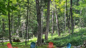 A forest clearing with Adirondack chairs and a fire pit. - Winter ‘27 Seasonal Available-Dog Friendly Chalet in the Woods + Pool, Mt. Snow (Wilmington)