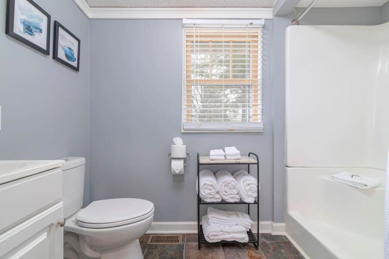 A bathroom with a toilet, a sink, a towel rack with folded towels, a window with blinds, and a shower area.