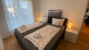 Una cama doble con ropa de cama estampada con motivos de estrellas, dos mesitas de noche con lámparas, un cuadro sobre la cama y una ventana con cortinas traslúcidas.