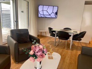 A modern living room with a sofa, a dining table with chairs, a flat-screen TV mounted on the wall, and a vase with flowers on a coffee table. - 2701 - Strandwiese 16 - Apartment 2701 in the Strandwiese (Dahme)