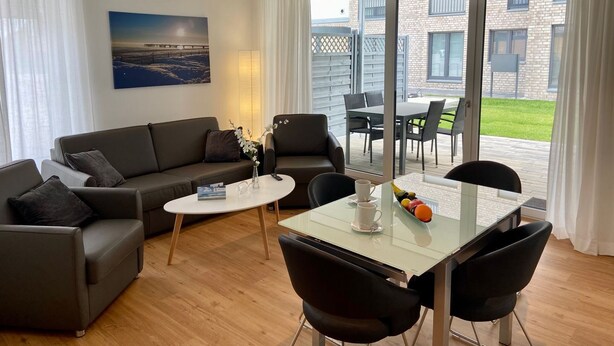 Una sala moderna con un sofá, una mesa de centro y un comedor. Hay un cuadro en la pared y vistas al exterior a través de amplias ventanas.