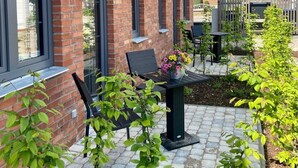 Een bakstenen huis met grote ramen, een terras met tafel en stoelen, en een tuin met planten.