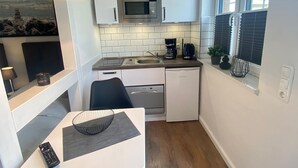 Een kleine keuken met witte kastjes, een magnetron, koffiezetapparaat en een spoelbak. Er staat een eettafel met stoelen en een afstandsbediening op.