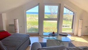 A modern living room with a large glass table, grey sofas, and a view of the sea through large windows.