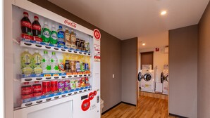 Máquina expendedora con diversas bebidas y aperitivos, lavadora y suelo de madera en una habitación de hotel.