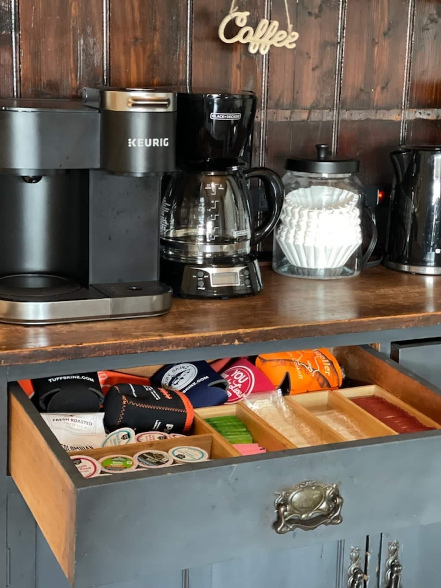 A coffee station with a Keurig machine, a glass carafe, and a coffee pot. Below, a drawer contains various coffee pods and a tea bag.