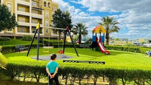 A playground with swings and a slide, surrounded by well-maintained hedges and a building in the background. - Charming beach apartment. Urb Canela Park (Playa de Isla Canela)