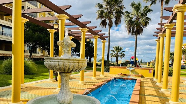 A courtyard with a central fountain, a swimming pool, and yellow columns. - Charming beach apartment. Urb Canela Park (Playa de Isla Canela)