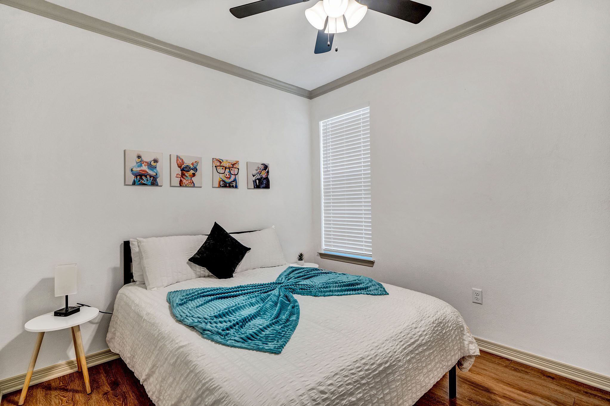 Quarto com cama, ventoinha no teto, janela com persianas e três quadros pendurados na parede.