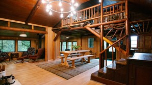 A rustic cabin interior with a wooden dining table, benches, a staircase, and a cozy seating area.