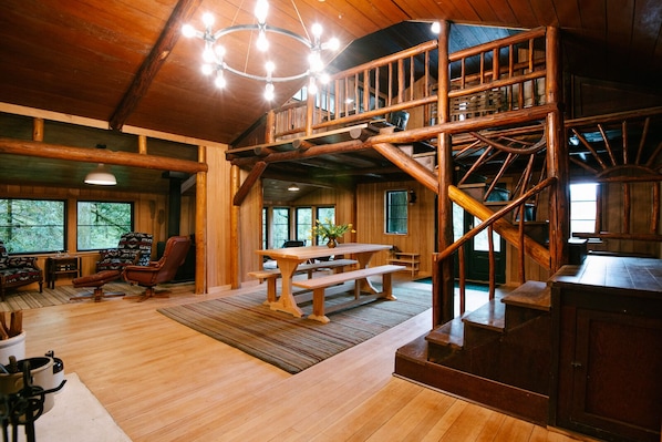 Interior rústico de uma cabana com mesa de jantar de madeira, bancos, escada e um espaço de estar aconchegante.