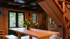 A wooden interior with a long table and benches, a vase with yellow flowers, and large windows with a view of trees.
