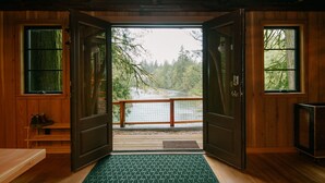 A wooden cabin with a large open door leading to a balcony overlooking a river and forest.