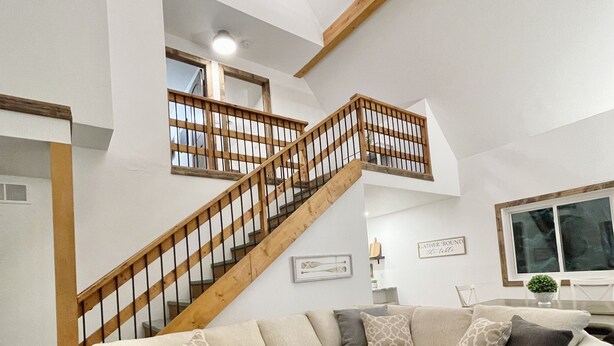 Ein geräumiges Wohnzimmer mit einem großen, L-förmigen beigen Sofa, einer Holztreppe mit Geländer und einem runden Ottomanen mit geometrischem Muster.