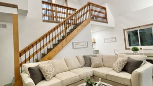 Ein Wohnzimmer mit einer Holztreppe, einem beigefarbenen Ecksofa und einem gemusterten Hocker.