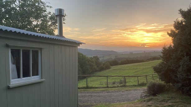 A small cabin with a window and a chimney, surrounded by a grassy landscape and a wooden fence. - Pet Friendly - 2 Guests - Shepherds Hut (Longhope)