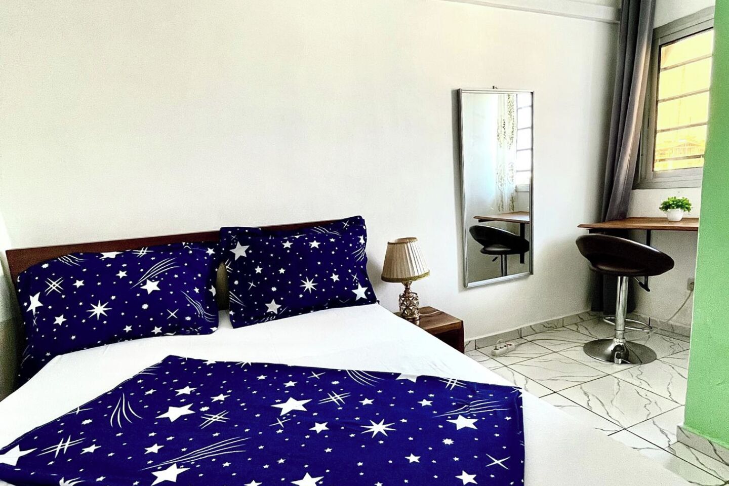 A bed with star-patterned bedding, a bedside lamp, a mirror, a desk with a chair, and a window with curtains.