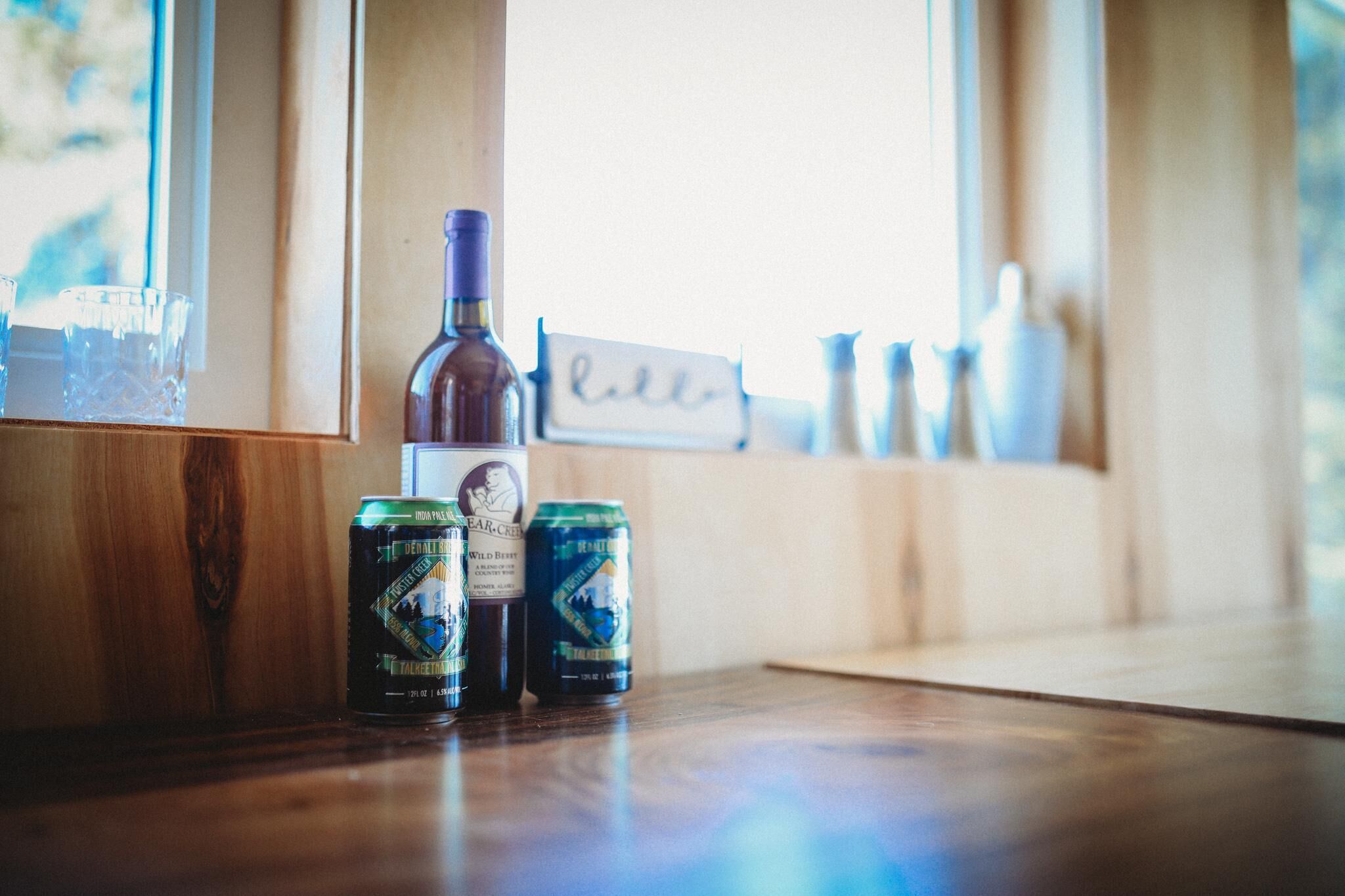 Une étagère en bois avec une bouteille et deux canettes de bière, un verre et une pancarte sur laquelle est écrit le mot « bonjour ».