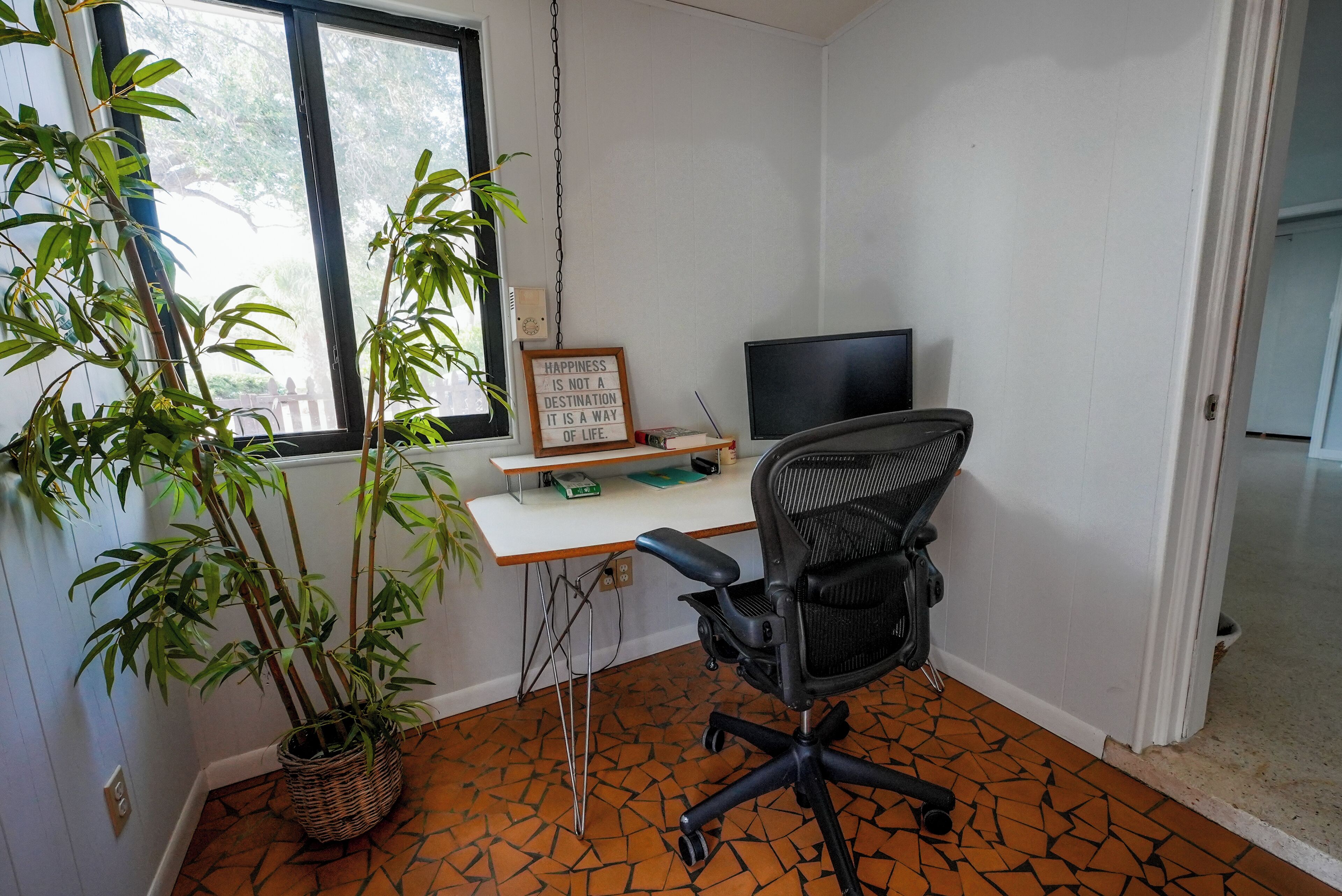 Quarto com escrivaninha, cadeira, plantas em vasos, televisão e uma frase motivacional na parede.