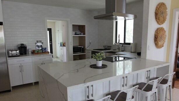 Eine moderne Küche mit weißen Schränken, einer großen Kochinsel mit Barhocker, einem eingebauten Backofen und einer Edelstahl-Dunstabzugshaube.