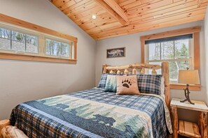 A cozy bedroom with a wooden ceiling, a large bed with a quilt, two windows, and a small bedside table with a lamp.