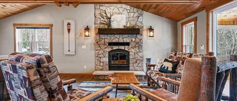A cozy living room with a stone fireplace, wooden furniture, and large windows.