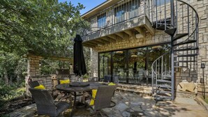 Patio esterno con tavolo in pietra e sedie in vimini, un barbecue e una zona relax coperta con una scala che conduce a un secondo piano.