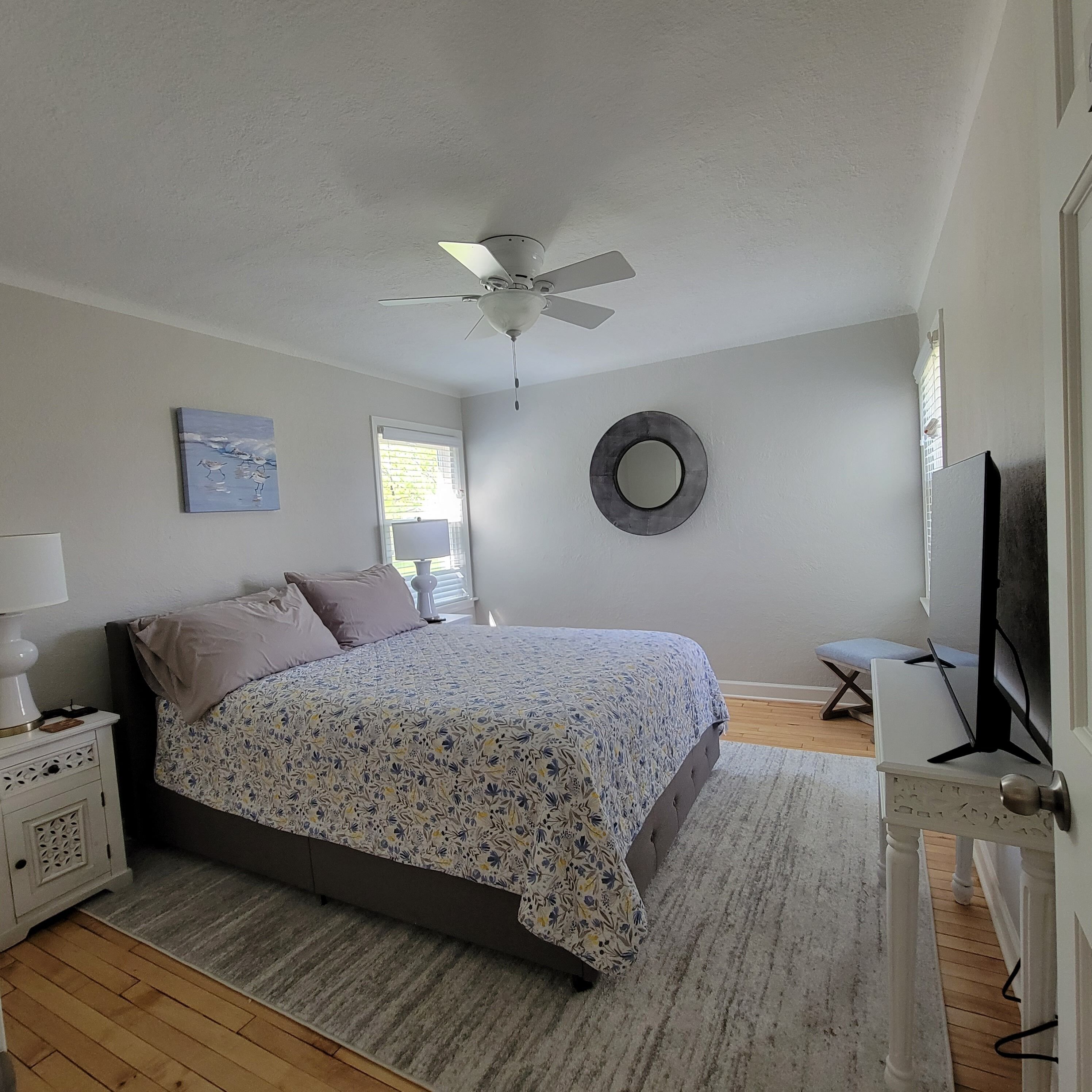 Une chambre à coucher avec un lit, un ventilateur de plafond, un miroir rond, une fenêtre et un téléviseur.