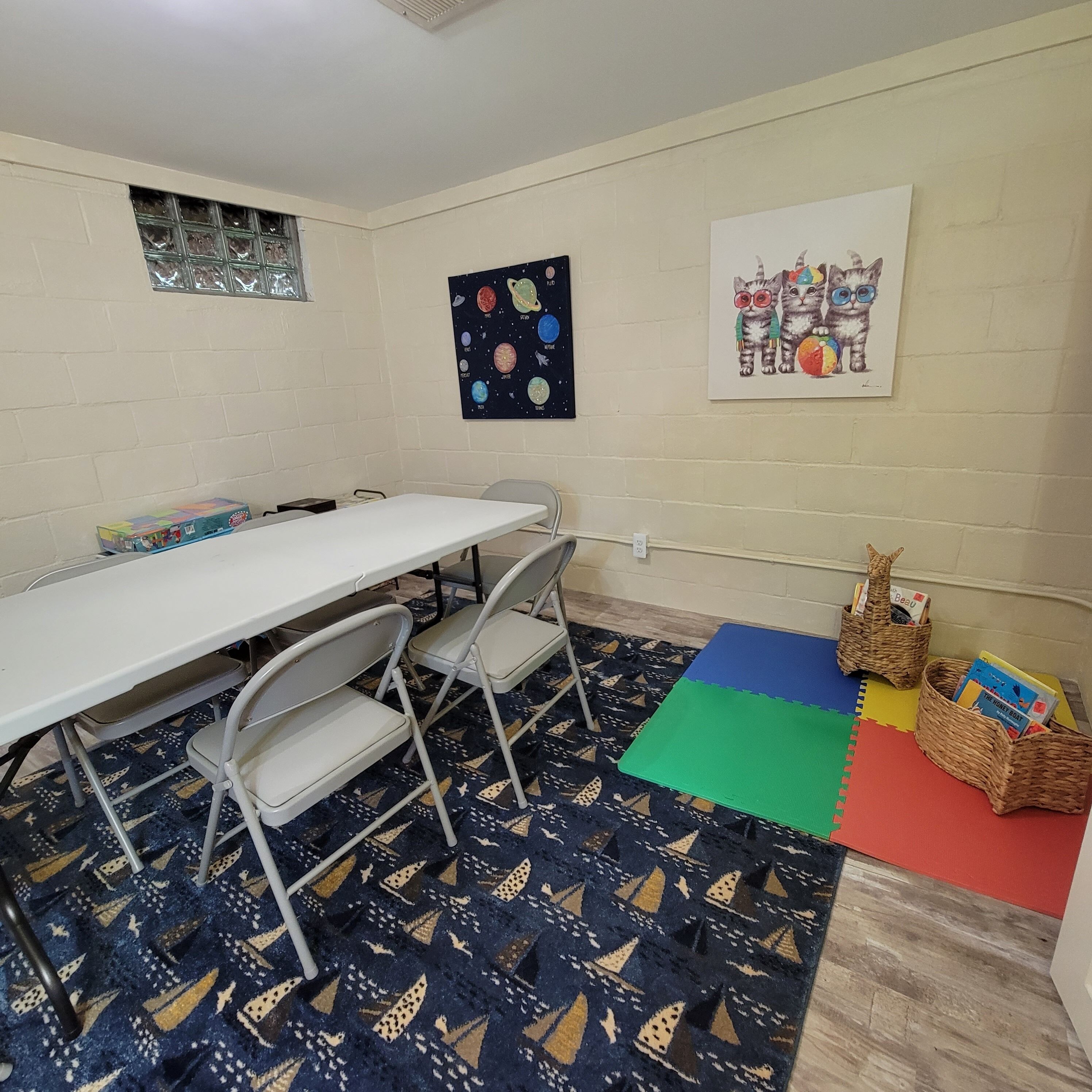 Een klaslokaal met een witte tafel, stoelen, een vloerkleed met een geometrisch patroon, een muur met een poster over ruimte en een lijst met foto's van kittens.