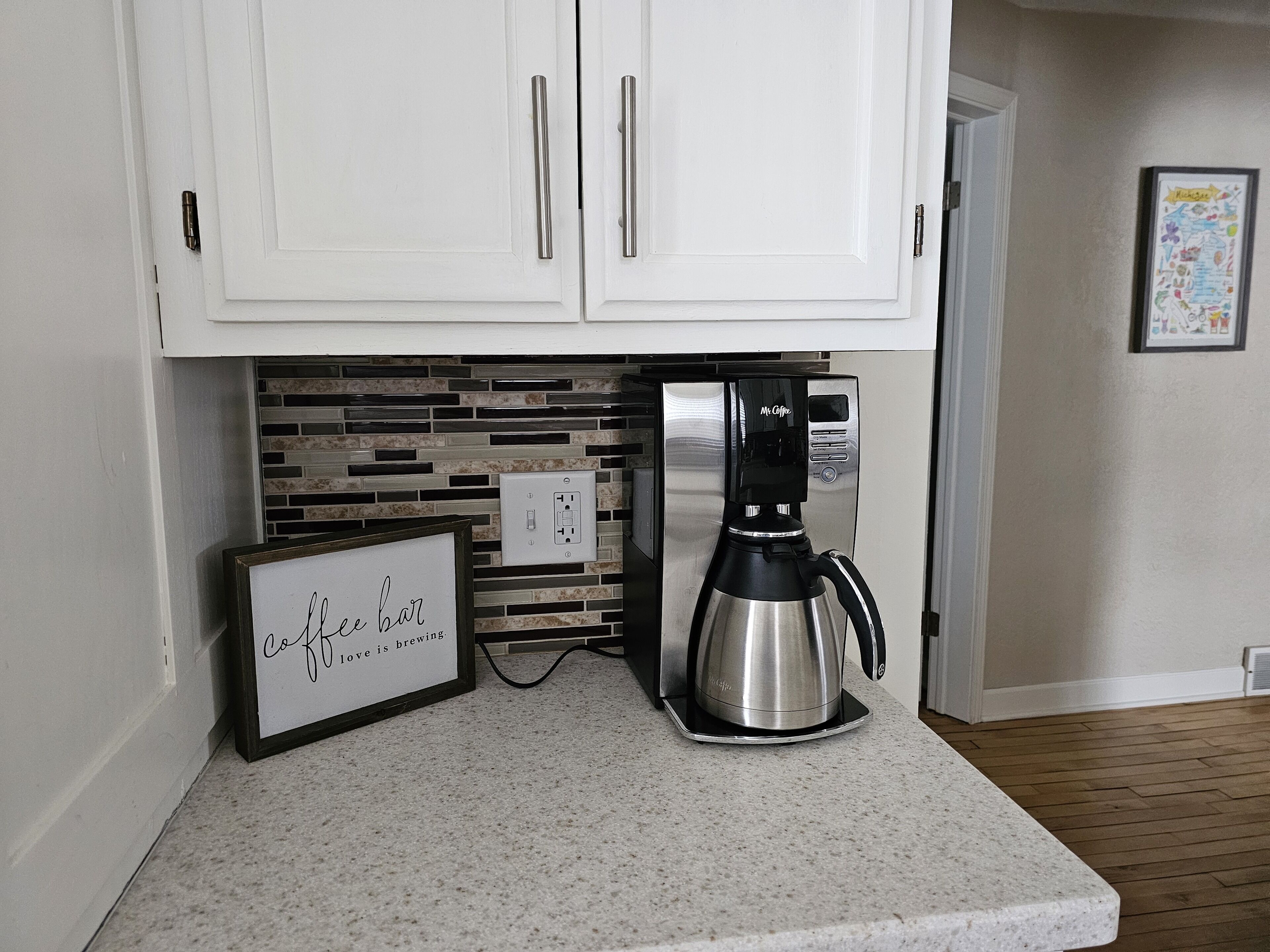 A kitchen with white cabinets, a coffee maker, and a framed sign that reads "coffee bar love is brewing".