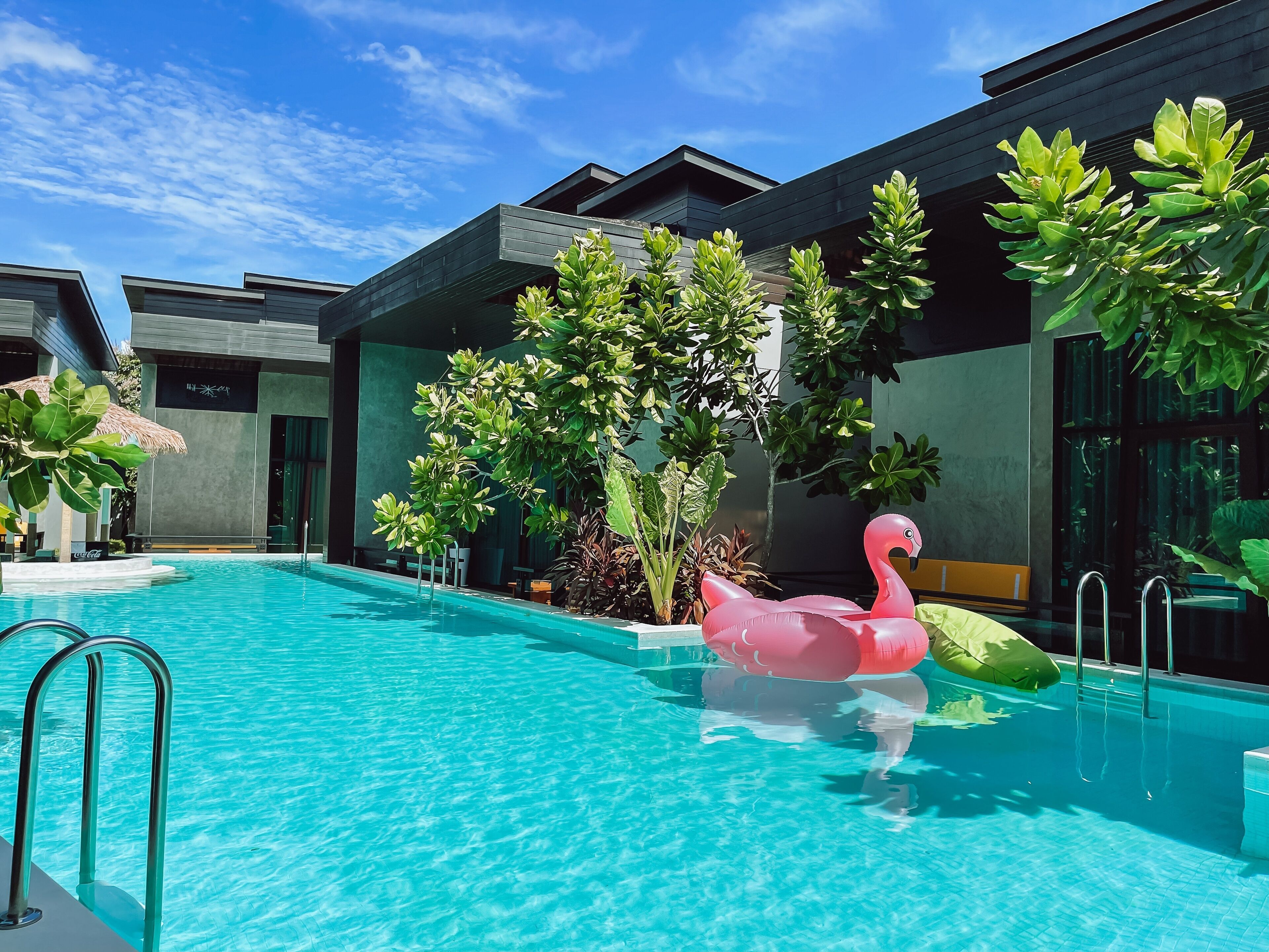 A swimming pool with a pink flamingo float, surrounded by modern buildings and lush greenery.
