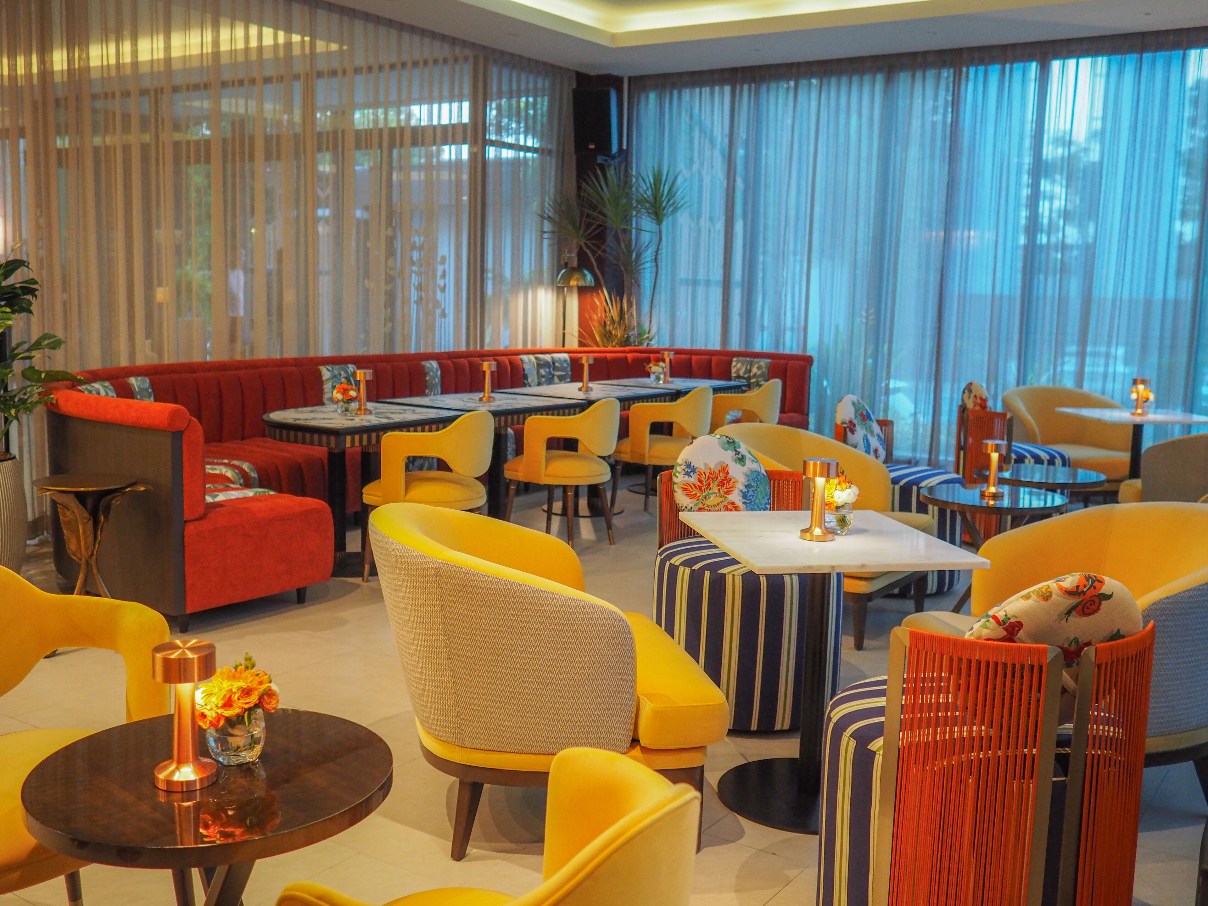 A modern hotel lobby with a mix of seating options including sofas and chairs, a central dining area with tables and chairs, and large windows with sheer curtains.