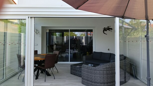 Eine moderne Terrasse mit Glastür zu einem Innenbereich, ausgestattet mit einem Esstisch, Stühlen und einer Rattan-Couch unter einem großen Sonnenschirm.