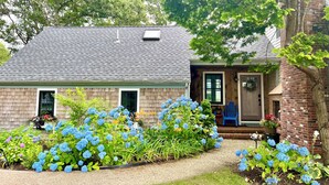 Una casa con facciata in mattoni, un portico con una panchina e un giardino con fiori di ortensie blu.